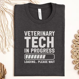 Puede incluir: Camiseta gris oscuro con el texto blanco "VETERINARY TECH IN PROGRESS", un gráfico de barra de carga y las palabras "LOADING... PLEASE WAIT". La camiseta está doblada y descansa sobre una superficie clara.