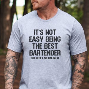 May include: A light grey t-shirt with the text "IT'S NOT EASY BEING THE BEST BARTENDER BUT HERE I AM NAILING IT" in black. The shirt is short-sleeved.