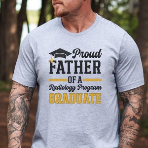 May include: A light gray t-shirt with the text "Proud FATHER of a Radiology Program GRADUATE" in black and gold. The design includes a graduation cap. The shirt is short-sleeved.