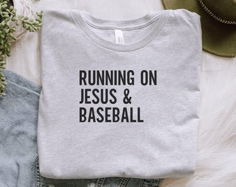 May include: A light gray crew neck t-shirt with the text "RUNNING ON JESUS & BASEBALL" in black block letters. The shirt is made of a soft material and is suitable for casual wear.