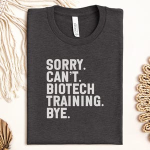 Puede incluir: Camiseta gris oscuro con el texto blanco "SORRY. CAN'T. BIOTECH TRAINING. BYE." La camiseta tiene cuello redondo y está hecha de un material suave. La camiseta está cuidadosamente doblada sobre una superficie blanca.