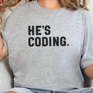 May include: Gray t-shirt with the text "HE'S CODING." printed in black.