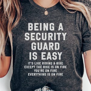Peut inclure: T-shirt gris foncé avec le texte "ÊTRE UN AGENT DE SÉCURITÉ EST FACILE." Le t-shirt contient également le texte "C'EST COMME FAIRE DU VÉLO SAUF QUE LE VÉLO EST EN FEU VOUS ÊTES EN FEU TOUT EST EN FEU."