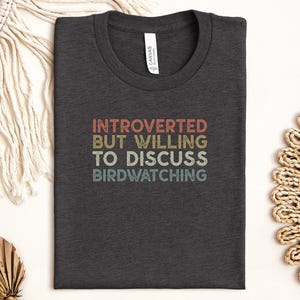 May include: Dark gray t-shirt with the text "INTROVERTED BUT WILLING TO DISCUSS BIRDWATCHING" in a retro font. The shirt is folded neatly, showcasing the design. A casual, comfortable tee for bird enthusiasts.