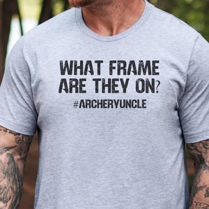 May include: A light grey t-shirt featuring the black text "WHAT FRAME ARE THEY ON?" and the hashtag "#ARCHERYUNCLE". The short-sleeved shirt is likely made of a soft fabric.