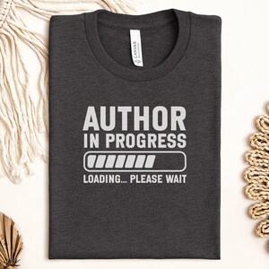 May include: Dark gray t-shirt with the text "AUTHOR IN PROGRESS" in white, along with a loading bar graphic and the words "LOADING... PLEASE WAIT". The shirt is folded and lies on a white surface.