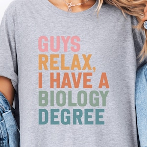 May include: A light gray t-shirt with the text "GUYS RELAX, I HAVE A BIOLOGY DEGREE" in pink, orange, green, and teal lettering. The shirt has rolled-up sleeves and is paired with a denim jacket.
