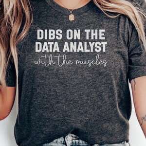 May include: A dark charcoal grey short-sleeved t-shirt featuring the white text DIBS ON THE DATA ANALYST with the muscles written in a combination of bold block lettering and a delicate cursive script across the centre of the chest.
