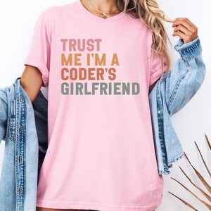 Pode incluir: Camiseta rosa claro com o texto "TRUST ME I'M A CODER'S GIRLFRIEND" em uma fonte retrô empilhada. A camisa é usada com uma jaqueta jeans.