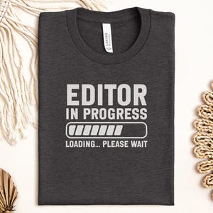 Peut inclure: T-shirt gris foncé avec l'inscription blanche « EDITOR IN PROGRESS ». En dessous, un indicateur de chargement et les mots « LOADING... PLEASE WAIT ». Le t-shirt plié est posé sur une surface blanche, avec des éléments décoratifs.