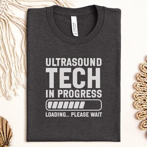 May include: Dark gray t-shirt with the words "ULTRASOUND TECH IN PROGRESS" in white block letters. Below the text is a loading bar graphic, and the words "LOADING... PLEASE WAIT" are printed below the graphic. The shirt is folded.
