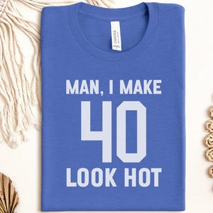 May include: A blue crew neck t-shirt with the white text "MAN, I MAKE 40 LOOK HOT". The number 40 is large and bold. The t-shirt is made of a soft material and is displayed flat on a white surface.