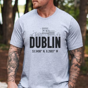 May include: A light gray t-shirt featuring the word "DUBLIN" in bold black letters, with a line drawing of a cityscape above it. Below the word "DUBLIN" are the coordinates 53.3498° N, 6.2603° W.