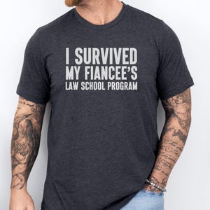 May include: Dark gray t-shirt with the text "I SURVIVED MY FIANCÉE'S LAW SCHOOL PROGRAM" in white block letters. The shirt is a crew neck style and is being worn by a person with tattoos on their arms.