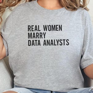May include: A light grey t-shirt featuring the black text "REAL WOMEN MARRY DATA ANALYSTS". The shirt is a relaxed fit, ideal for casual wear.