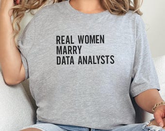 May include: A light gray t-shirt with the text "REAL WOMEN MARRY DATA ANALYSTS" in black block letters. The shirt is a casual style, suitable for everyday wear.