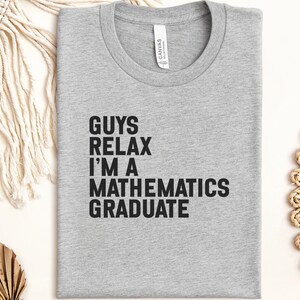 May include: A light grey t-shirt with the text "GUYS RELAX I'M A MATHEMATICS GRADUATE" in bold black letters. The shirt is folded neatly and has a crew neck. The shirt is made of a soft material. The shirt is a great gift for a mathematics graduate.