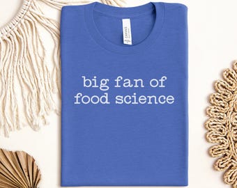 May include: A blue t-shirt with the text "big fan of food science" in white. The shirt is folded and lies flat on a neutral surface. The shirt has a crew neck and a tag that says "CANVAS".