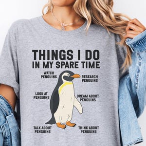 May include: A heather gray t-shirt featuring a central graphic of a penguin. The text reads: THINGS I DO IN MY SPARE TIME, WATCH PENGUINS, RESEARCH PENGUINS, LOOK AT PENGUINS, DREAM ABOUT PENGUINS, TALK ABOUT PENGUINS, and THINK ABOUT PENGUINS.