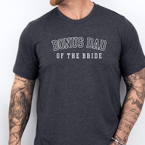 May include: Dark gray t-shirt with the text "BONUS DAD OF THE BRIDE" in white, outlined letters. The shirt has short sleeves and a crew neck. The person wearing the shirt has tattoos on their arms.