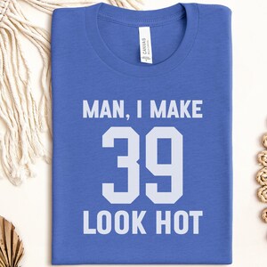May include: A blue t-shirt with the text "MAN, I MAKE 39 LOOK HOT" in white block letters. The shirt is folded and has a tag that says "CANVAS". The shirt is surrounded by decorative items.