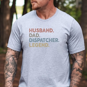 May include: A light gray t-shirt with the words "HUSBAND. DAD. DISPATCHER. LEGEND." in a retro font. The text is in red, blue, and yellow. The shirt is short-sleeved.