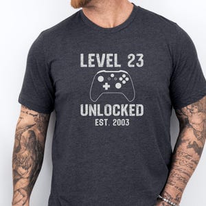 Könnte beinhalten: Dunkelgraues T-Shirt mit dem Text "LEVEL 23 UNLOCKED EST. 2003" und einer weißen Controller-Grafik. Das T-Shirt hat einen Rundhalsausschnitt und ist aus weichem Material. Ein tolles Geschenk für einen Gamer.