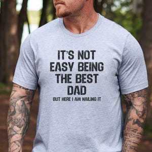 May include: A gray t-shirt with the text "It's not easy being the best dad, but here I am nailing it."