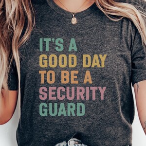 May include: Dark gray t-shirt with the text "IT'S A GOOD DAY TO BE A SECURITY GUARD" in colorful block letters. The shirt is a crew neck and has short sleeves. A gold necklace is visible.