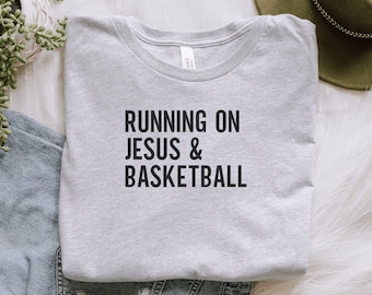 May include: A light gray t-shirt with the text "RUNNING ON JESUS & BASKETBALL" in black block letters. The shirt is made of a soft material and has a crew neck. The shirt is folded on a white surface.