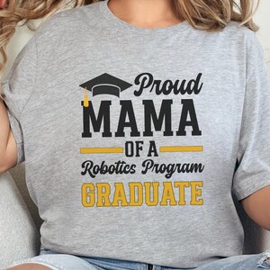 May include: A light gray t-shirt with the text "Proud MAMA of a Robotics Program GRADUATE" in black and gold. The design includes a graduation cap graphic. The shirt is a casual style.