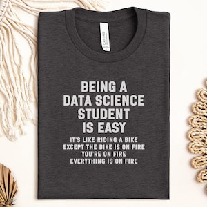 May include: Dark gray t-shirt with white text that reads "BEING A DATA SCIENCE STUDENT IS EASY." The text continues with "IT'S LIKE RIDING A BIKE EXCEPT THE BIKE IS ON FIRE YOU'RE ON FIRE EVERYTHING IS ON FIRE."