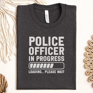 May include: Dark gray t-shirt with the text "POLICE OFFICER IN PROGRESS" in white, along with a loading bar graphic and the words "LOADING... PLEASE WAIT". The shirt is folded and has a tag that says "CANVAS".