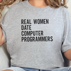 May include: A light grey t-shirt featuring the black text "REAL WOMEN DATE COMPUTER PROGRAMMERS". The shirt has a relaxed fit, ideal for casual wear.