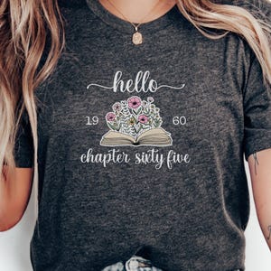 May include: Dark gray t-shirt with the text "hello" above an open book illustration with flowers and the text "19 60 chapter sixty five". The shirt has a crew neck.
