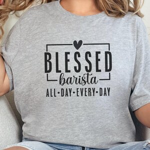 May include: A light gray t-shirt with the words "BLESSED barista ALL-DAY EVERY-DAY" in black. The text is inside a black rectangular frame with a heart above the word "BLESSED". The shirt is casual wear.