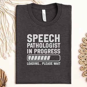 Könnte beinhalten: Dunkelgraues T-Shirt mit weißem Text "SPEECH PATHOLOGIST IN PROGRESS", einem Ladebalken-Grafik und den Worten "LOADING... PLEASE WAIT". Das T-Shirt ist gefaltet und hat ein "CANVAS"-Etikett.