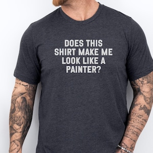 May include: Dark gray t-shirt with the text "DOES THIS SHIRT MAKE ME LOOK LIKE A PAINTER?" in white. The shirt is short-sleeved and has a crew neck. The person wearing the shirt has tattoos on their arms.