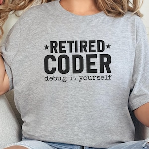 Könnte beinhalten: Hellgraues T-Shirt mit dem schwarzen Aufdruck "RETIRED CODER debug it yourself". Der Text ist zentriert, mit "RETIRED" über "CODER". Das T-Shirt hat einen lässigen Stil und ist bequem.
