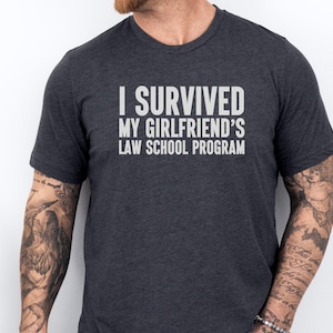 May include: A dark gray t-shirt with the text "I survived my girlfriend's law school program" printed in white.