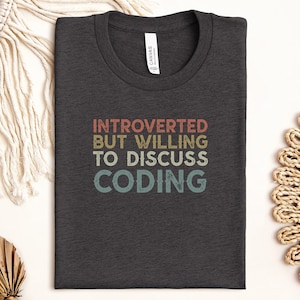 May include: Dark gray t-shirt with the text "INTROVERTED BUT WILLING TO DISCUSS CODING" in a retro font. The shirt is folded and has a tag on the collar. The shirt is surrounded by decorative items.