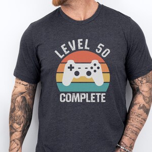 May include: Dark gray t-shirt with a retro design featuring a white game controller and the text "LEVEL 50 COMPLETE." The design is set against a colorful sunset-style background. A casual tee for gamers.