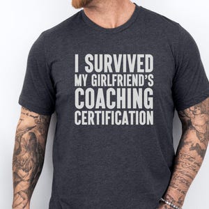 May include: A dark grey t-shirt with the text "I survived my girlfriend's coaching certification" printed in white.
