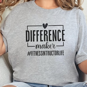 Peut inclure: T-shirt gris clair avec l'inscription noire "DIFFERENCE maker", un cœur et le hashtag "#FITNESSINSTRUCTORLIFE". Le t-shirt est de style décontracté, idéal pour les entraînements ou le quotidien.