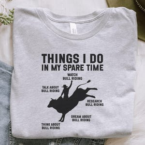 May include: A light gray t-shirt with the text "THINGS I DO IN MY SPARE TIME" and a graphic of a bull rider. The graphic is black and shows a cowboy riding a bucking bull. The shirt also lists activities related to bull riding.