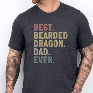 May include: Dark grey t-shirt featuring the text "BEST. BEARDED DRAGON. DAD. EVER." in a vintage, distressed font. The text is in shades of red, yellow, green, and grey. The shirt has a crew neck.