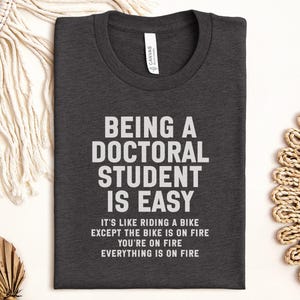 May include: Charcoal grey t-shirt with white text: "BEING A DOCTORAL STUDENT IS EASY". The shirt also features the phrases: "IT'S LIKE RIDING A BIKE EXCEPT THE BIKE IS ON FIRE YOU'RE ON FIRE EVERYTHING IS ON FIRE".