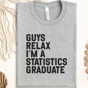 May include: A grey t-shirt with the text "GUYS RELAX I'M A STATISTICS GRADUATE" in bold black letters. The shirt is folded neatly, with a tag visible at the neckline. The shirt is a crew neck style.