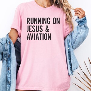 May include: A light pink t-shirt with the text "RUNNING ON JESUS & AVIATION" in black block letters. The shirt has short sleeves and a crew neck. A denim jacket is draped over the shoulders.