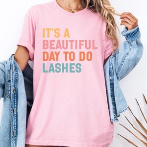 Könnte beinhalten: Hellrosa T-Shirt mit dem Text "IT'S A BEAUTIFUL DAY TO DO LASHES" in Orange, Rosa und Türkis. Das Shirt wird mit einer blauen Jeansjacke getragen. Das Thema ist Schönheit und Selbstpflege.